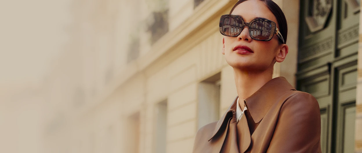 Woman wearing large sunglasses stands in front of a building and looks confidently into the distance. (photo)