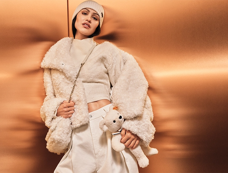 Woman in light winter clothing and a beanie leans against a metallic wall while holding a small plush toy. (photo)